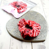 Red and white paisley scrunchie placed on a heart-shaped stone, with a pink packaging bag in view.