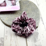Plum-colored scrunchie with a paisley design, set on a rustic heart-shaped stone with a pink plastic bag in the backdrop.