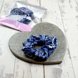 Navy scrunchie featuring a classic paisley print, on a stone heart with its packaging partially visible.