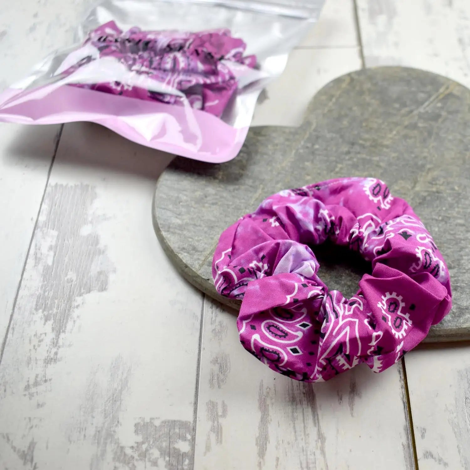 Vibrant pink paisley-patterned scrunchie displayed on a stone heart, with a clear plastic bag alongside.