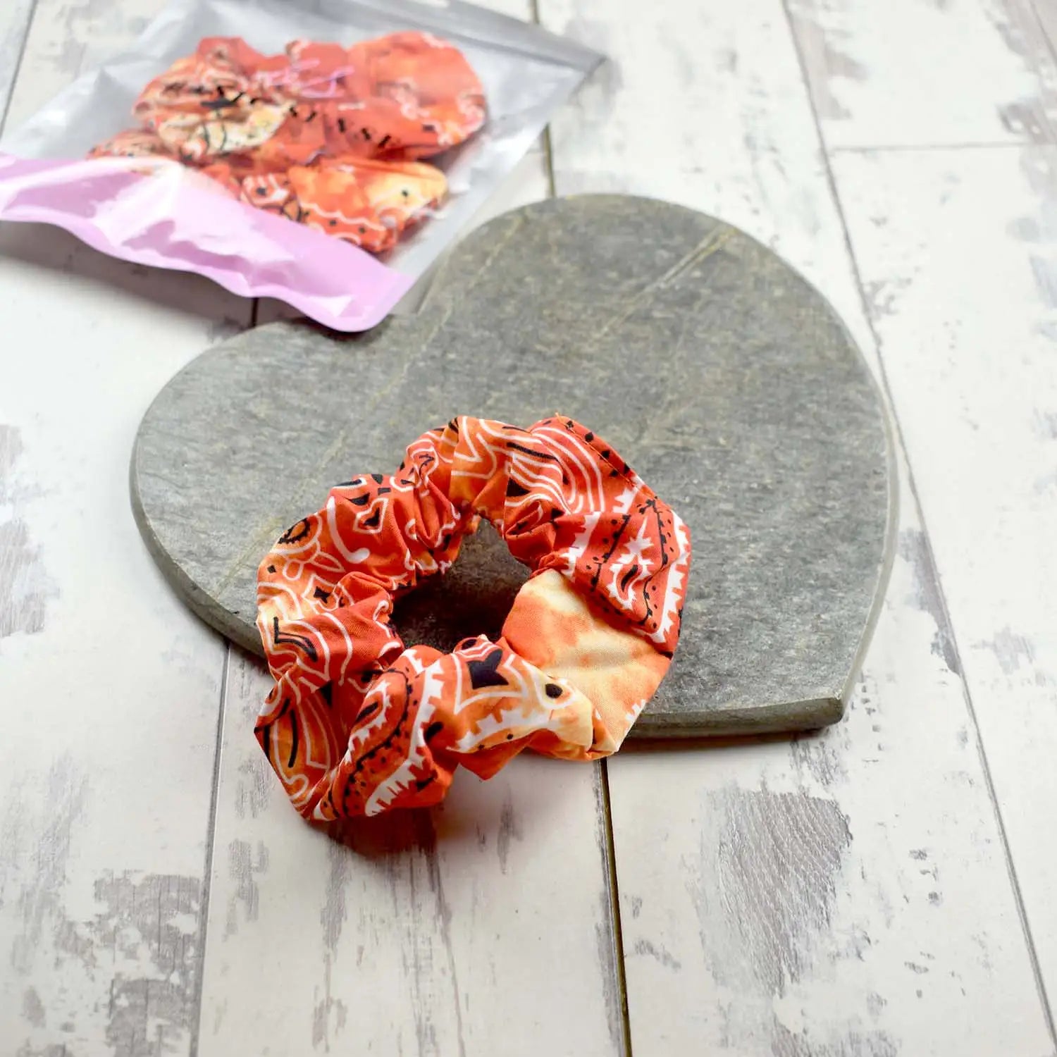 Orange scrunchie with red paisley accents, resting on a stone heart, includes packaging for retail display.