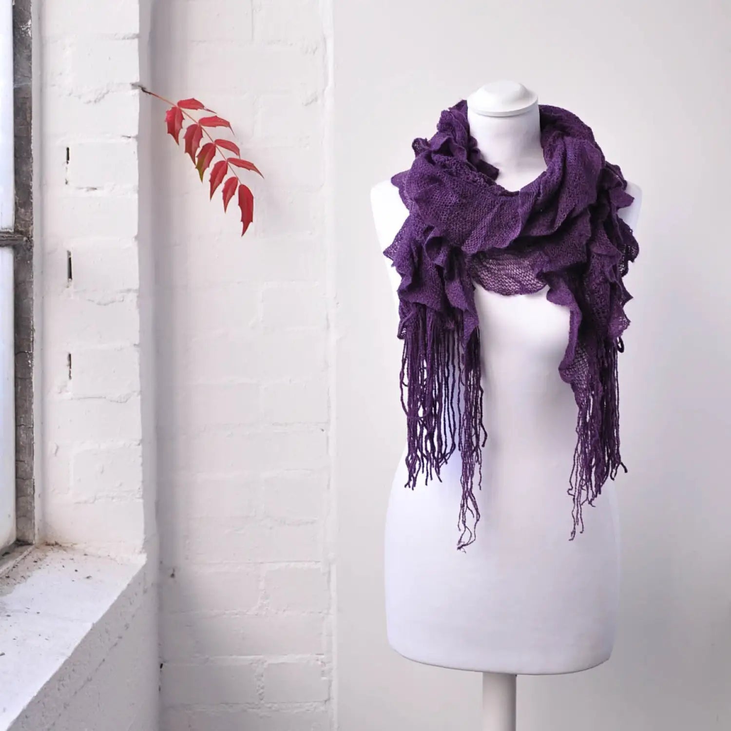 Lightweight knitted ruffled purple scarf on mannequin dummy in front of white brick wall